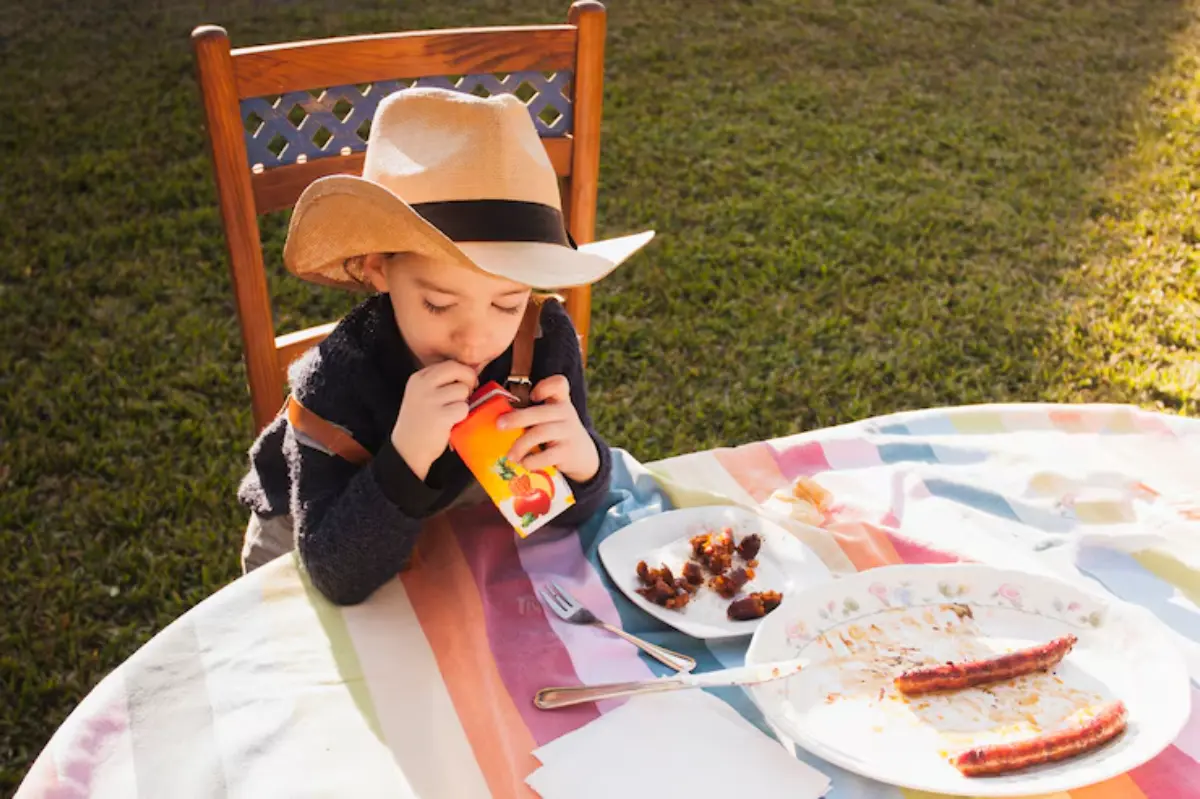 A child in a cowboy hat sits at a table with colorful tablecloth, holding a juice box, surrounded by empty plates and leftover food.