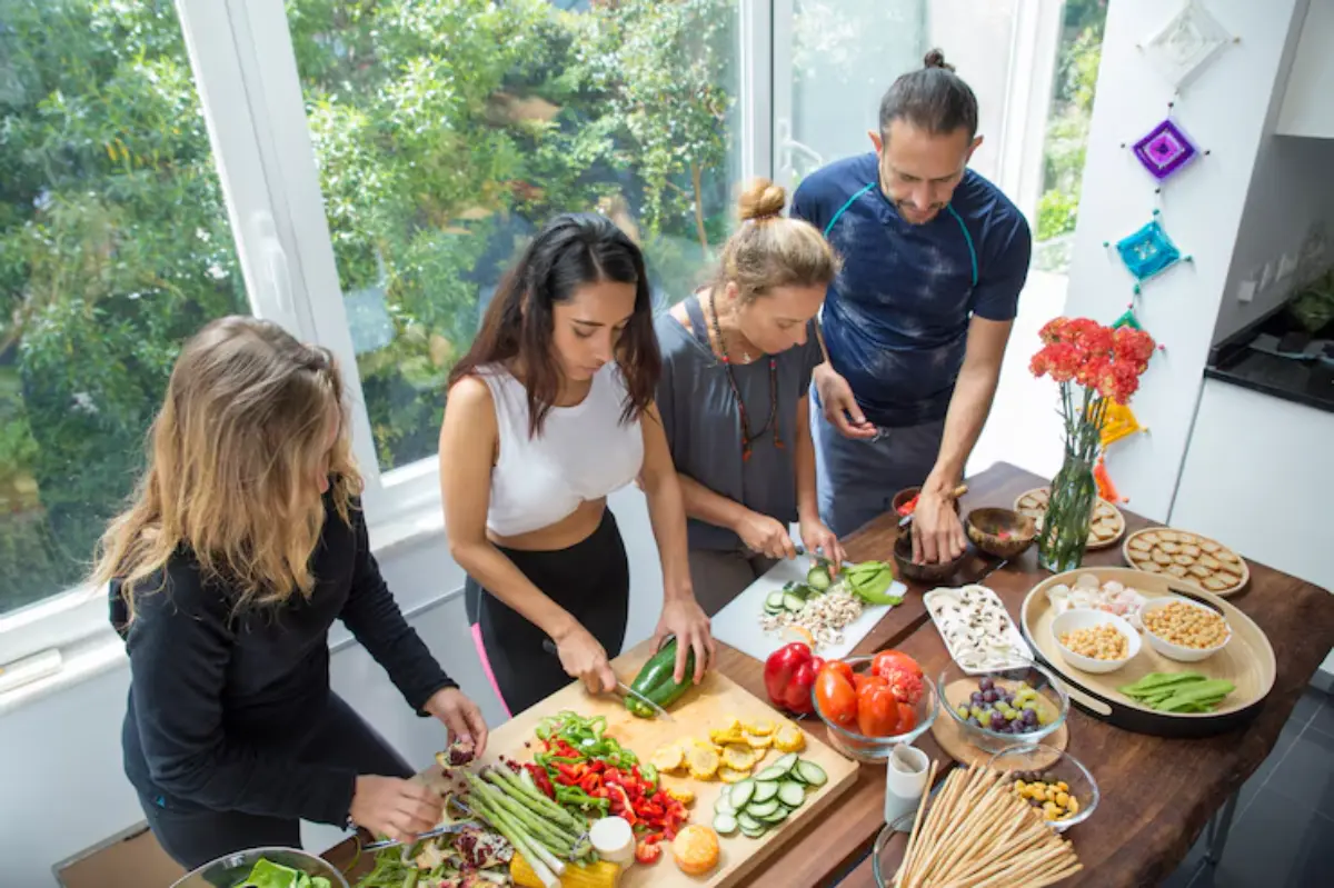 A group of four people preparing colorful vegetables and fruit on a wooden table in a bright kitchen, with various dishes displayed.