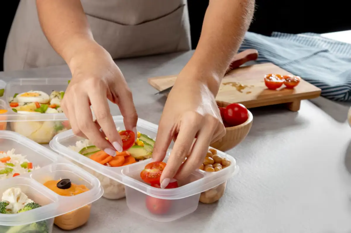 A person arranging sliced tomatoes on colorful meal prep containers filled with various fresh ingredients on a kitchen countertop.