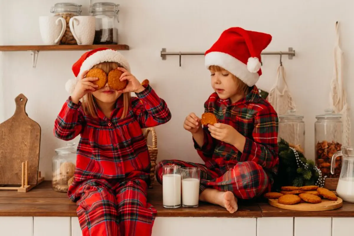 Two children in festive red plaid pajamas and Santa hats enjoy cookies and milk on a wooden counter decorated for the holidays.