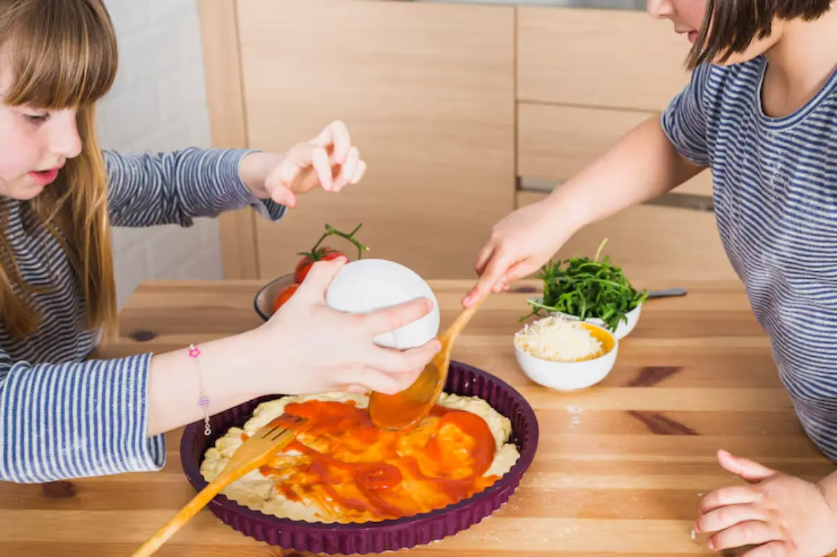 Two children are preparing pizza together, spreading sauce on dough, surrounded by fresh ingredients and toppings on a wooden table.