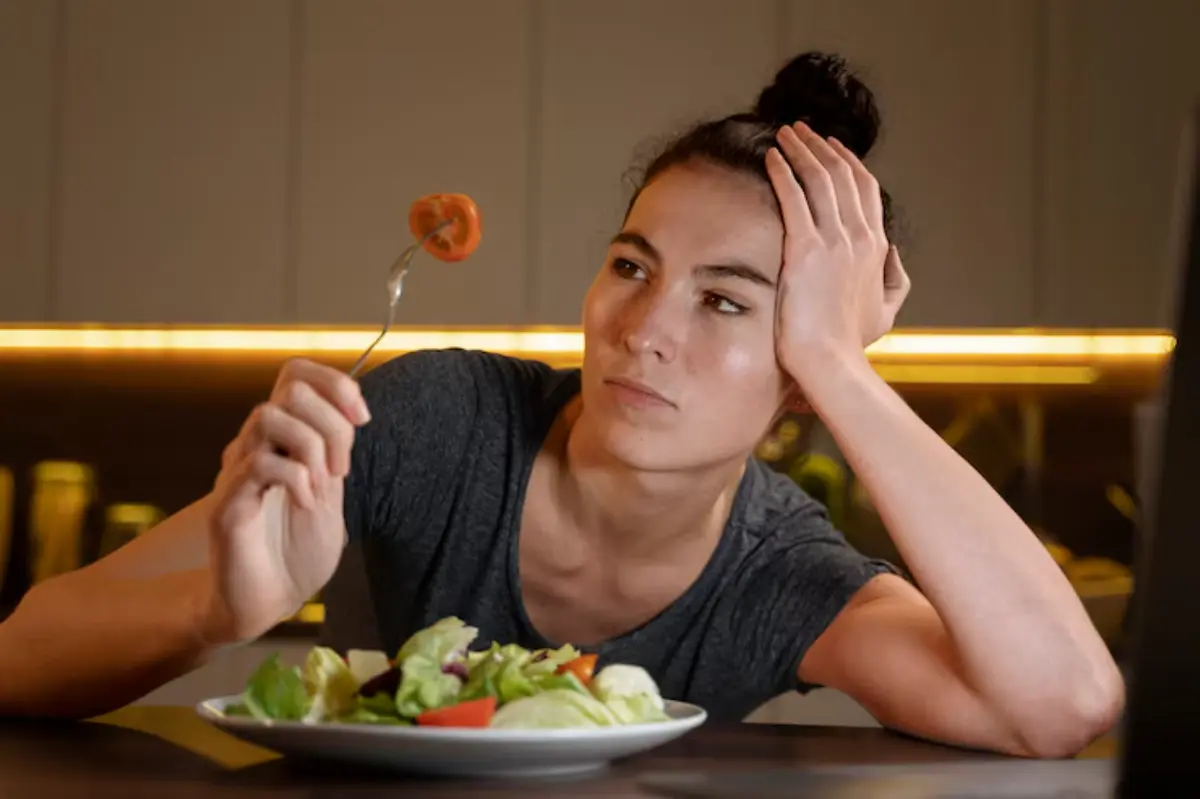 A person sits at a table holding a fork with a piece of tomato, looking contemplatively at a salad on a plate in front of them.