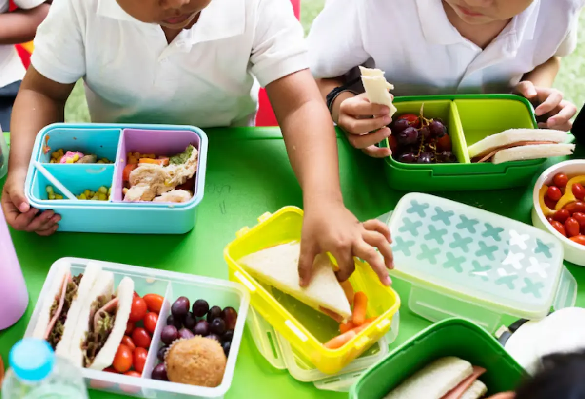 Two children at a green table enjoy colorful lunchboxes filled with sandwiches, fruits, and vegetables, sharing their meals together.
