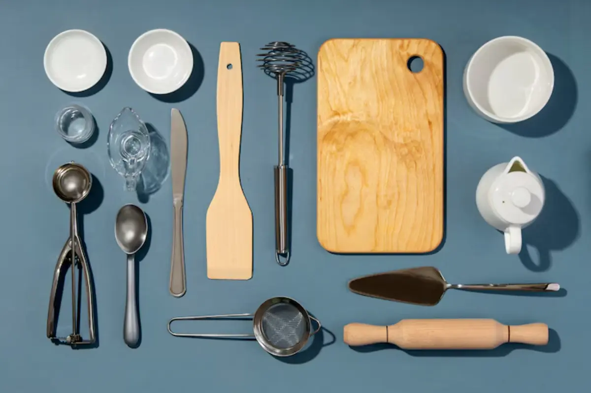 A flat lay of various kitchen tools including spoons, measuring cups, a spatula, a whisk, and a wooden cutting board on a blue background.