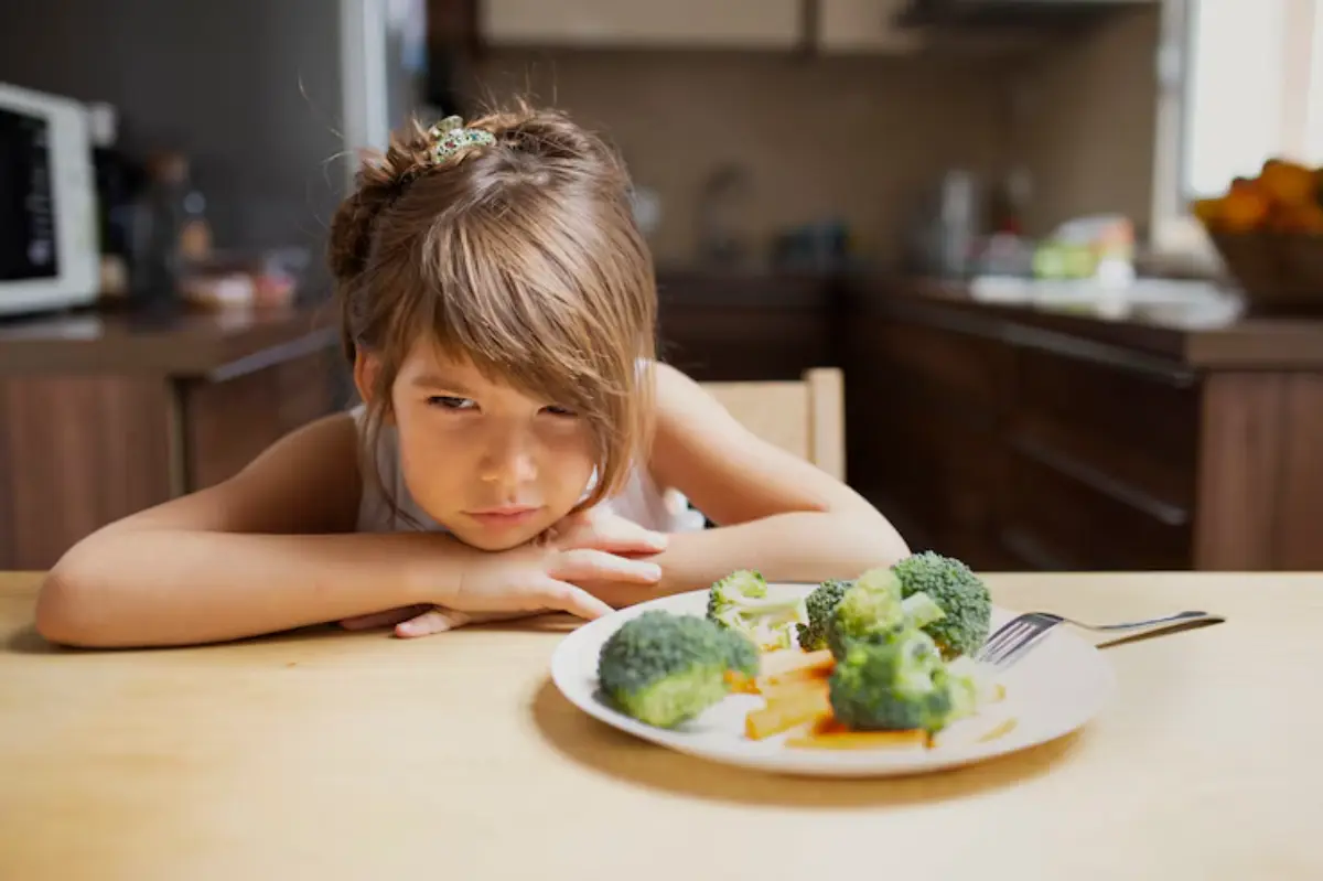 A child resting her head on the table, looking at a plate of broccoli and carrots in a cozy kitchen setting.