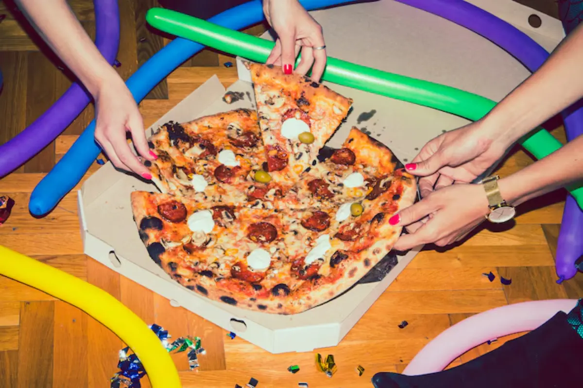 A group shares a large pizza with various toppings, surrounded by colorful party decorations on a wooden floor.