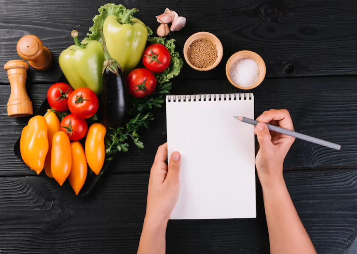 Hands holding a pencil over a blank notepad, surrounded by fresh vegetables and spices on a wooden table.