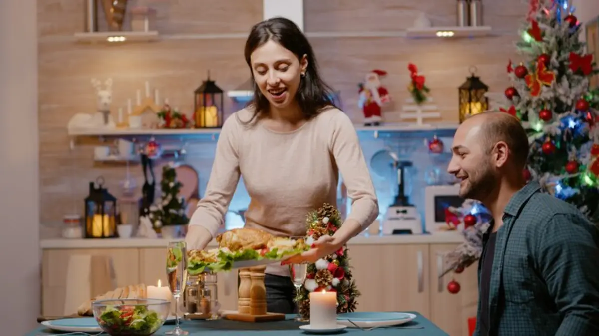 A woman serves a festive meal on a dining table adorned with candles and a Christmas tree, creating a cozy holiday atmosphere.