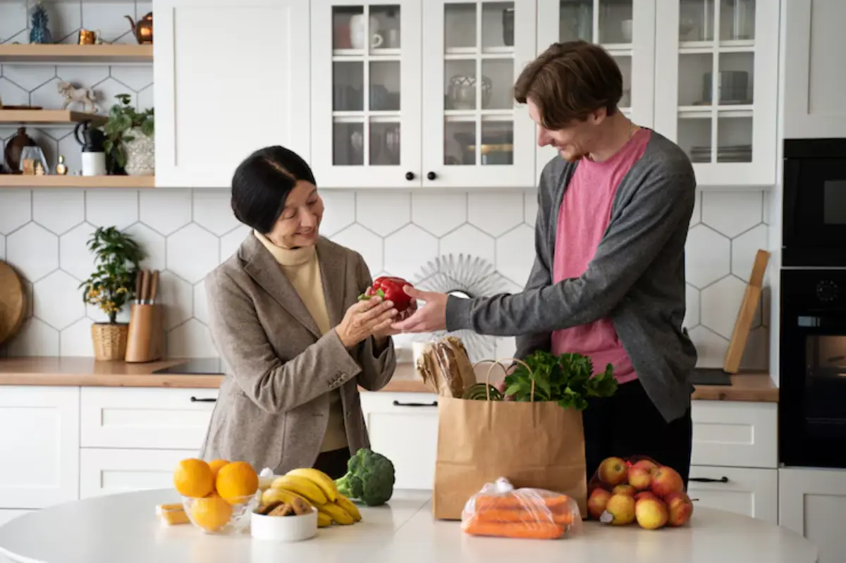 A woman receives a red bell pepper from a man in a kitchen, surrounded by fresh fruits and vegetables on a table.
