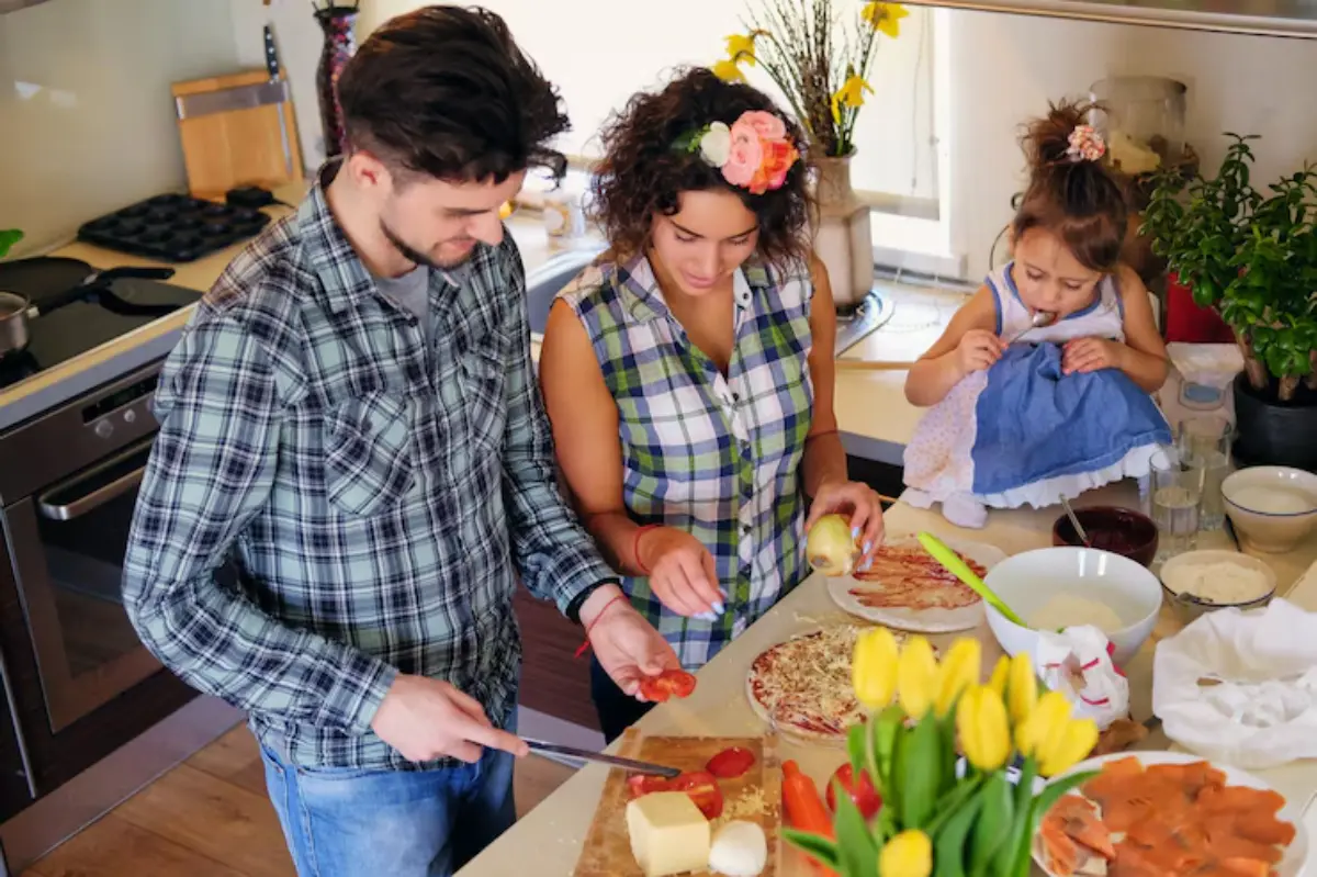 A family prepares food in a bright kitchen, with ingredients like cheese, tomatoes, and fresh flowers on the countertop.