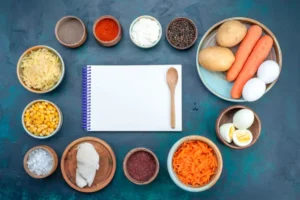 A flat lay of fresh ingredients: shredded cheese, corn, spices, vegetables, and eggs arranged around a blank notepad and wooden spoon.
