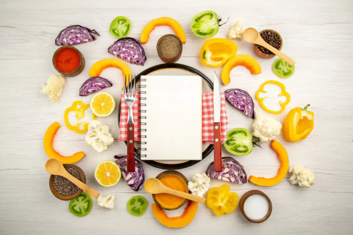 A blank notepad surrounded by colorful vegetables, spices, and utensils, creating a vibrant, culinary-themed flat lay.