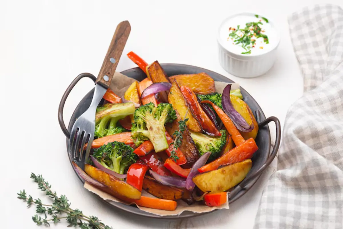 A plate of colorful roasted vegetables, including broccoli, carrots, and bell peppers, with a side of creamy dip and fresh herbs.