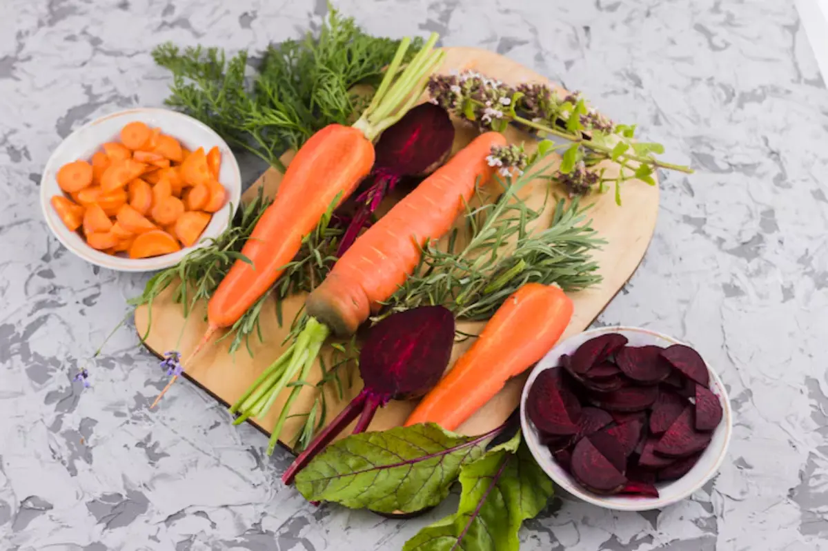 Fresh carrots and beets arranged on a wooden board with sliced carrots and beets in bowls, surrounded by herbs and greens.