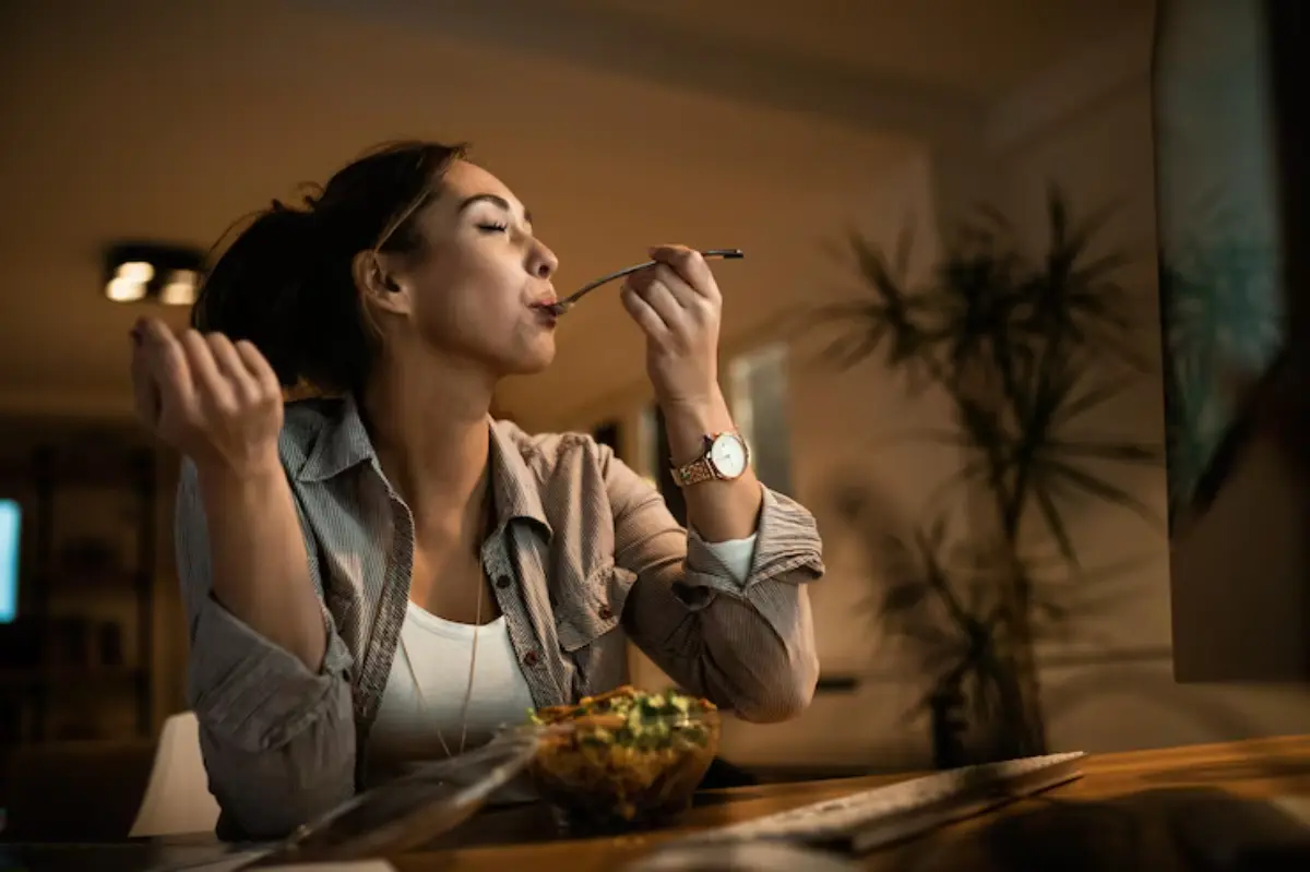 A woman seated at a table enjoys a meal from a clear bowl, with a cozy ambiance and a TV in the background.
