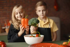 Two children hold vegetables—one with a red bell pepper and the other with broccoli—posing in a kitchen setting with a bowl of mixed veggies.