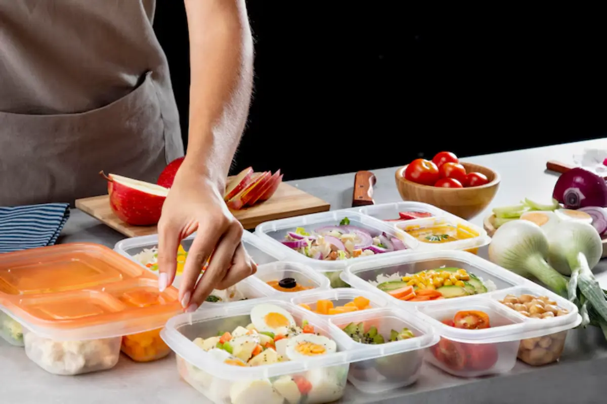 A person prepares healthy meal prep containers filled with fresh vegetables, eggs, and fruits on a kitchen countertop.