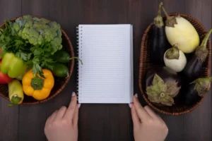 A hand holds an open notebook surrounded by baskets of fresh vegetables, including peppers, broccoli, and eggplants, on a wooden table.