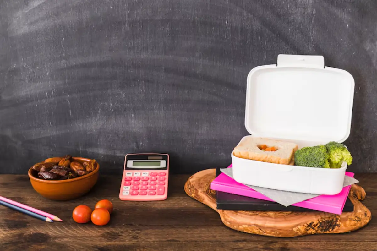 A white lunchbox filled with a sandwich and broccoli sits atop colorful notebooks beside a bowl of dates, cherry tomatoes, and a calculator.