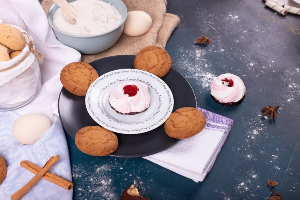 Freshly baked cookies and a cupcake with pink frosting sit on a black plate, surrounded by flour, spices, and eggs.