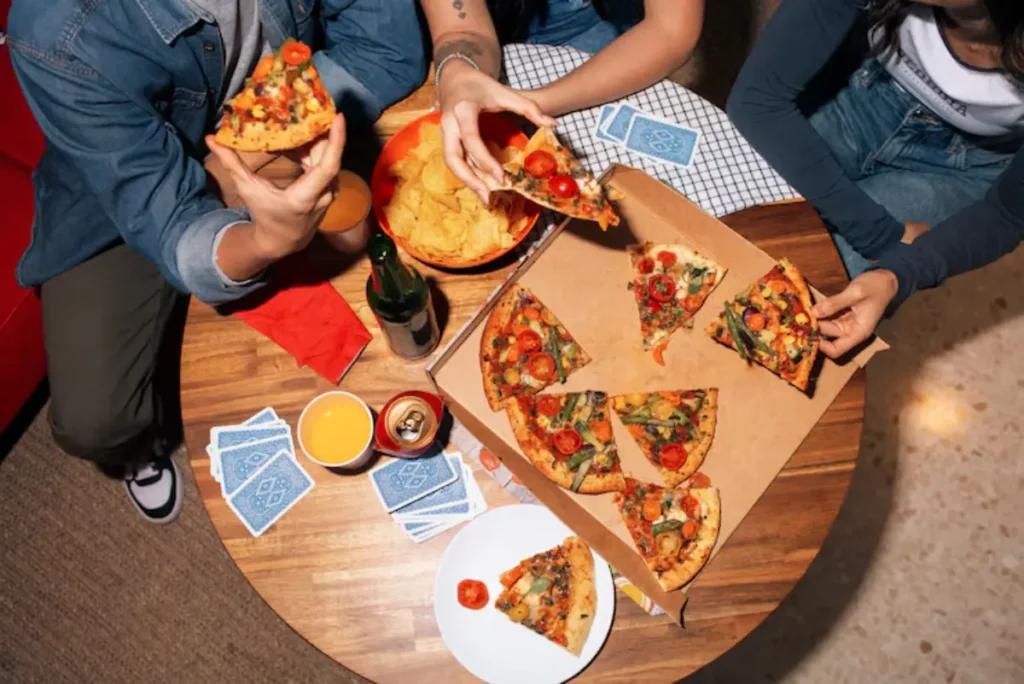 A group enjoying pizza and snacks around a wooden table, with slices and drinks, with colorful cards scattered nearby.