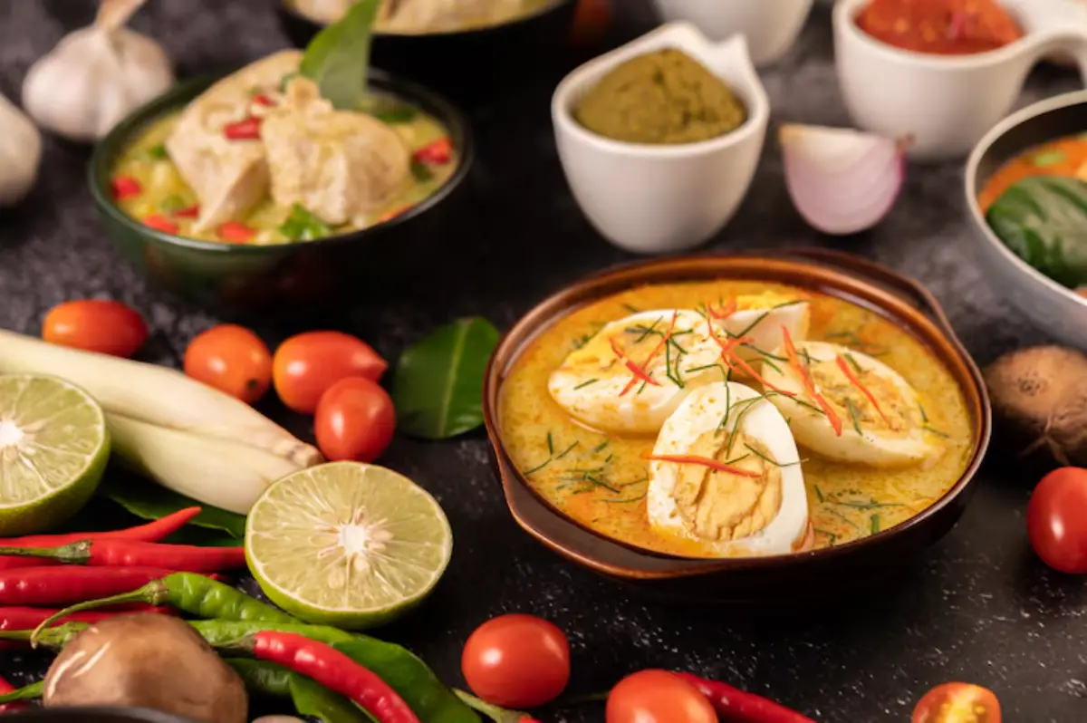 A flavorful dish of curry with sliced eggs, garnished with herbs, surrounded by fresh vegetables and spices on a dark background.