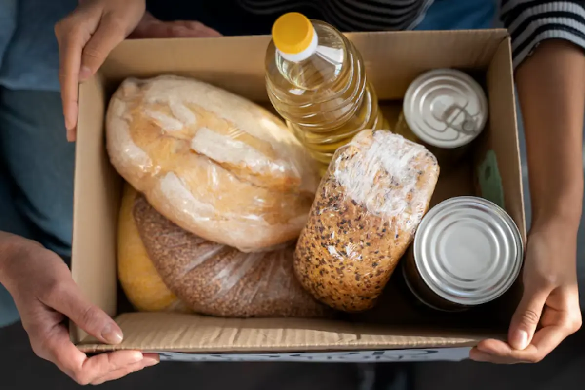 A person holds a cardboard box filled with various food items, including loaves of bread, cooking oil, and canned goods.