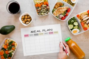 A meal prep scene featuring a weekly meal plan, colorful healthy dishes, an avocado, a drink, and a hand holding a pen.