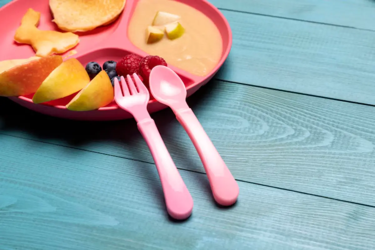 A pink divided plate filled with pancakes, fruit slices, and yogurt, accompanied by pink toddler utensils on a teal wooden table.