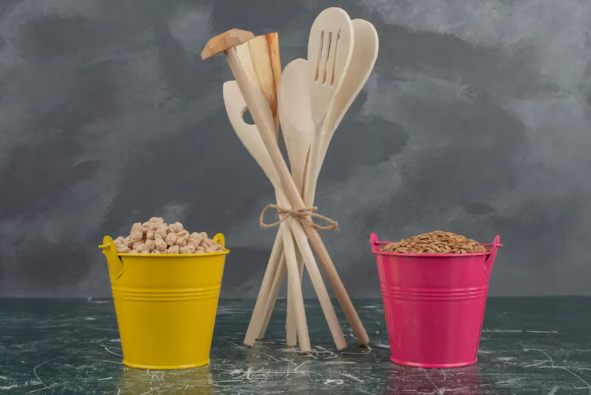 A yellow bucket filled with popcorn and a pink bucket with grains, next to wooden cooking utensils, against a dark textured background.