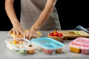 A person in an apron organizes colorful meal prep containers on a countertop, surrounded by fresh vegetables and a wooden bowl of tomatoes.