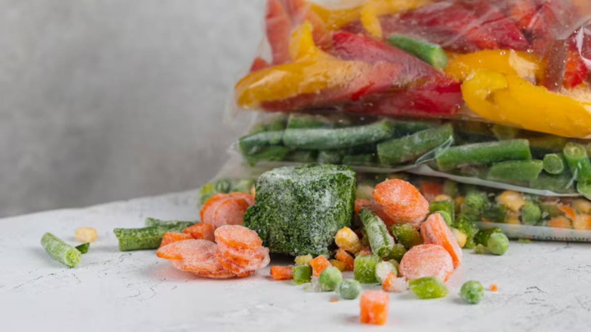 A clear bag of colorful frozen bell peppers sits behind scattered mixed frozen vegetables, including carrots, peas, and green beans.
