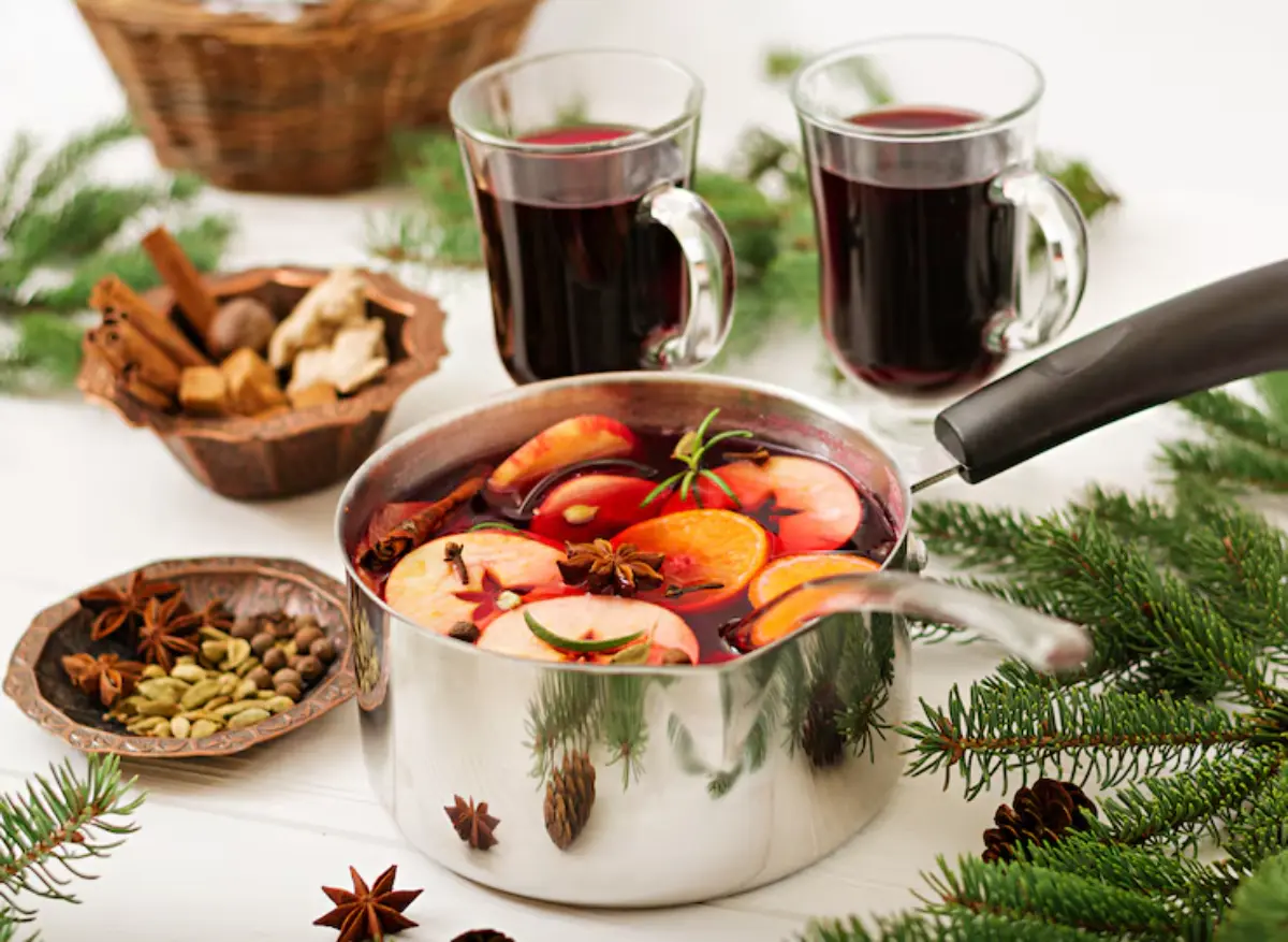 A pot of spiced fruit drink with apple and orange slices, surrounded by herbs, spices, and two mugs of beverage on a festive table.