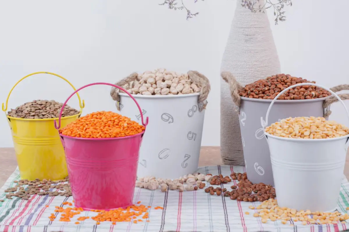 Colorful buckets filled with various legumes and grains are displayed on a checkered cloth, accompanied by a decorative vase.