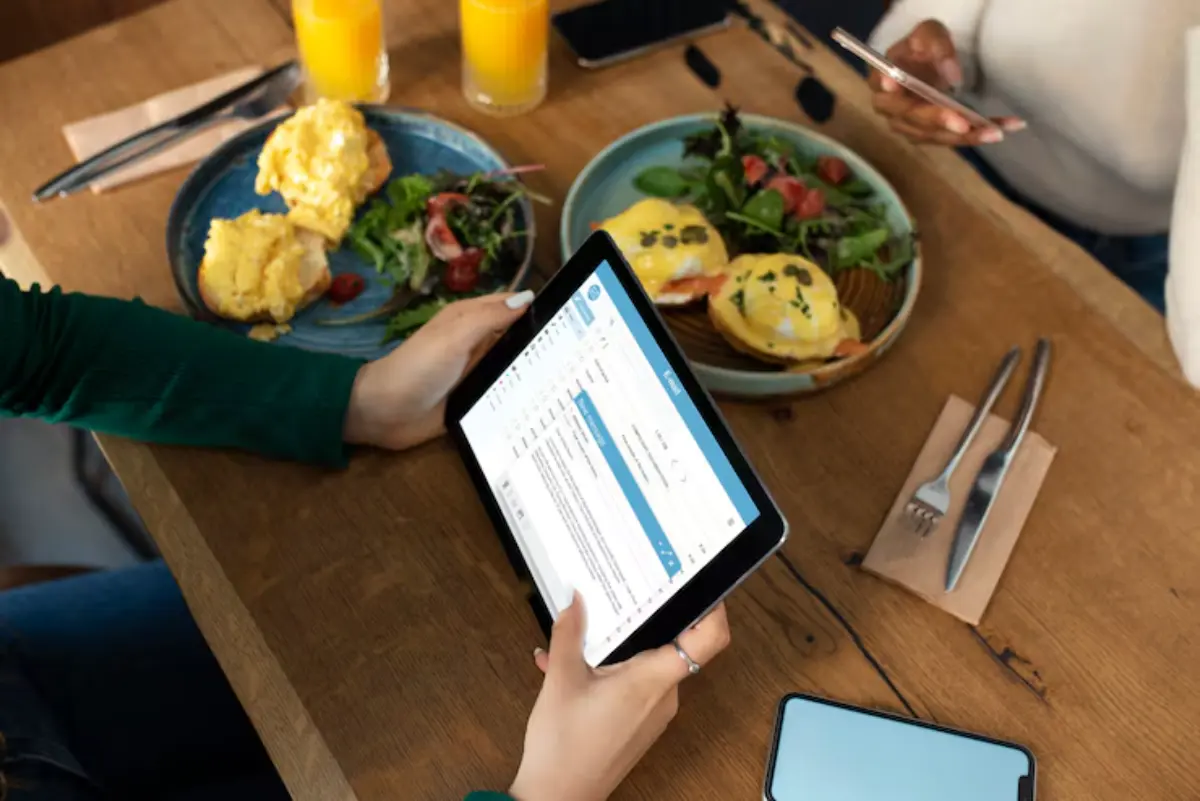 A person holds a tablet displaying text while seated at a table with brunch plates and orange juice, another person uses a phone nearby.