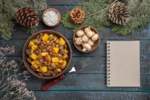 A wooden bowl of sautéed mushrooms and diced potatoes, surrounded by pinecones, herbs, and a blank notepad on a rustic table.
