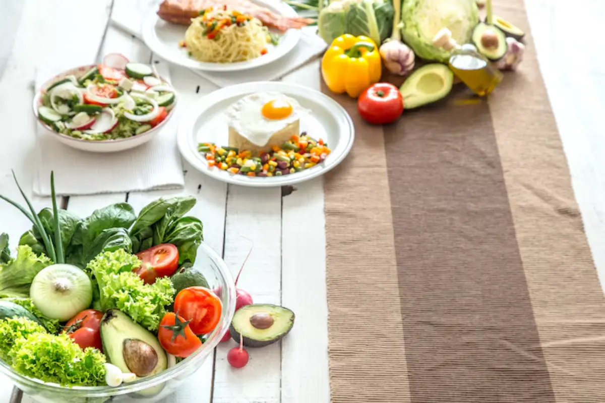 A variety of colorful, fresh dishes and vegetables arranged on a wooden table, including salads, pasta, and cooked vegetables.