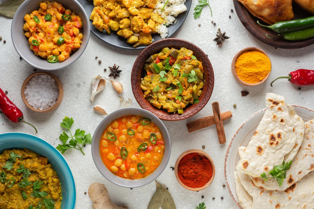 An array of vibrant Indian dishes, including curries, rice, and flatbreads, surrounded by spices and fresh ingredients on a textured surface.