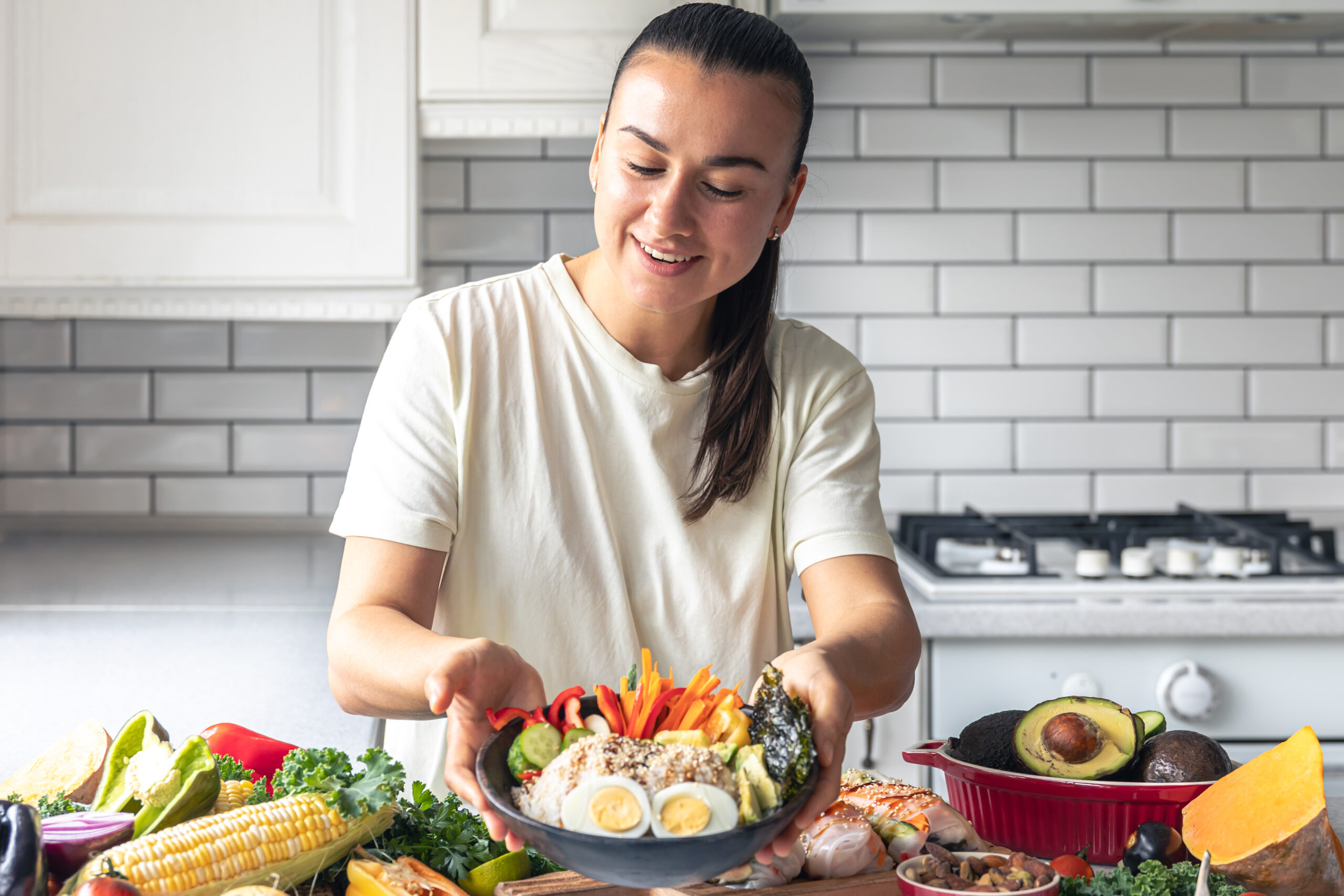 Young woman in a kitchen holding a plate with vegetables, rice, and eggs, smiling at the camera.